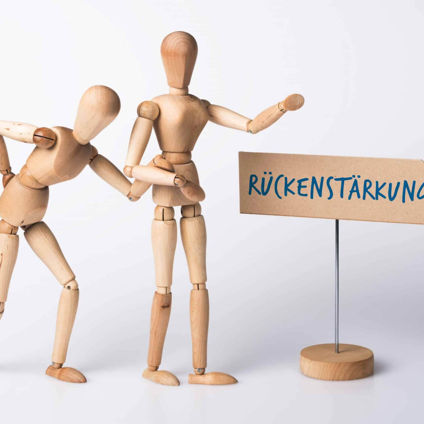 Zwei Holzfiguren, die vor einem Schild stehen, auf dem Rückenstärkung steht. Eine der beiden Holzfiguren hält sich mit einer Hand den unteren Rücken und mit der anderen Hand an der anderen Holzfigur fest.
